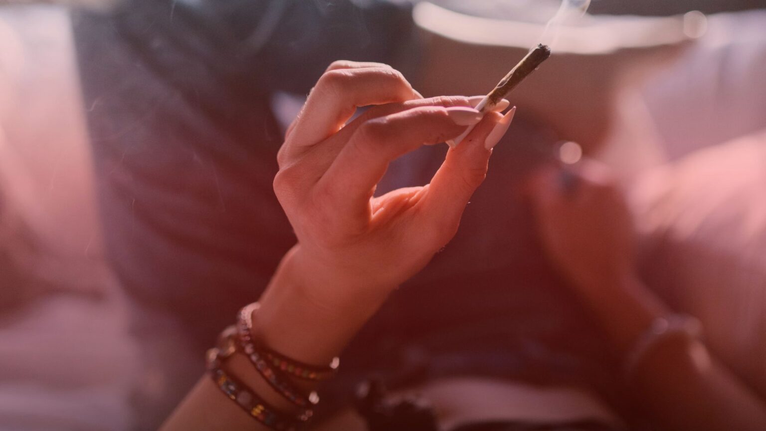 Nature's Canopy House A hand holding a smoking rolled cigarette, with an arm adorned with beaded bracelets. Blurry background of a person's legs and a couch or bed. Dispensary In East York and York