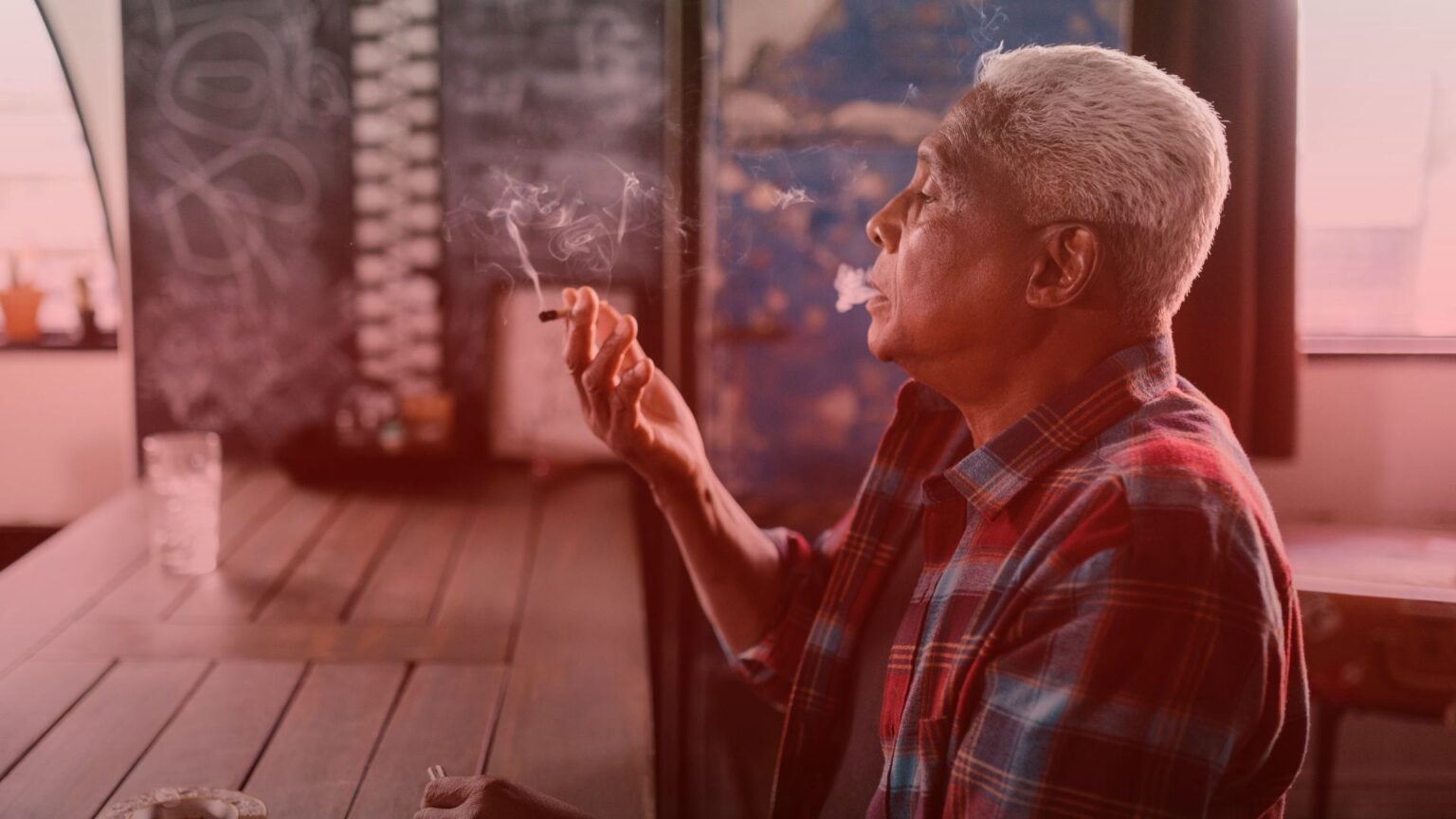 Nature's Canopy House An older man with short white hair and a red plaid shirt smokes a cigarette while sitting at a wooden table in a room with dim lighting. Dispensary In East York and York