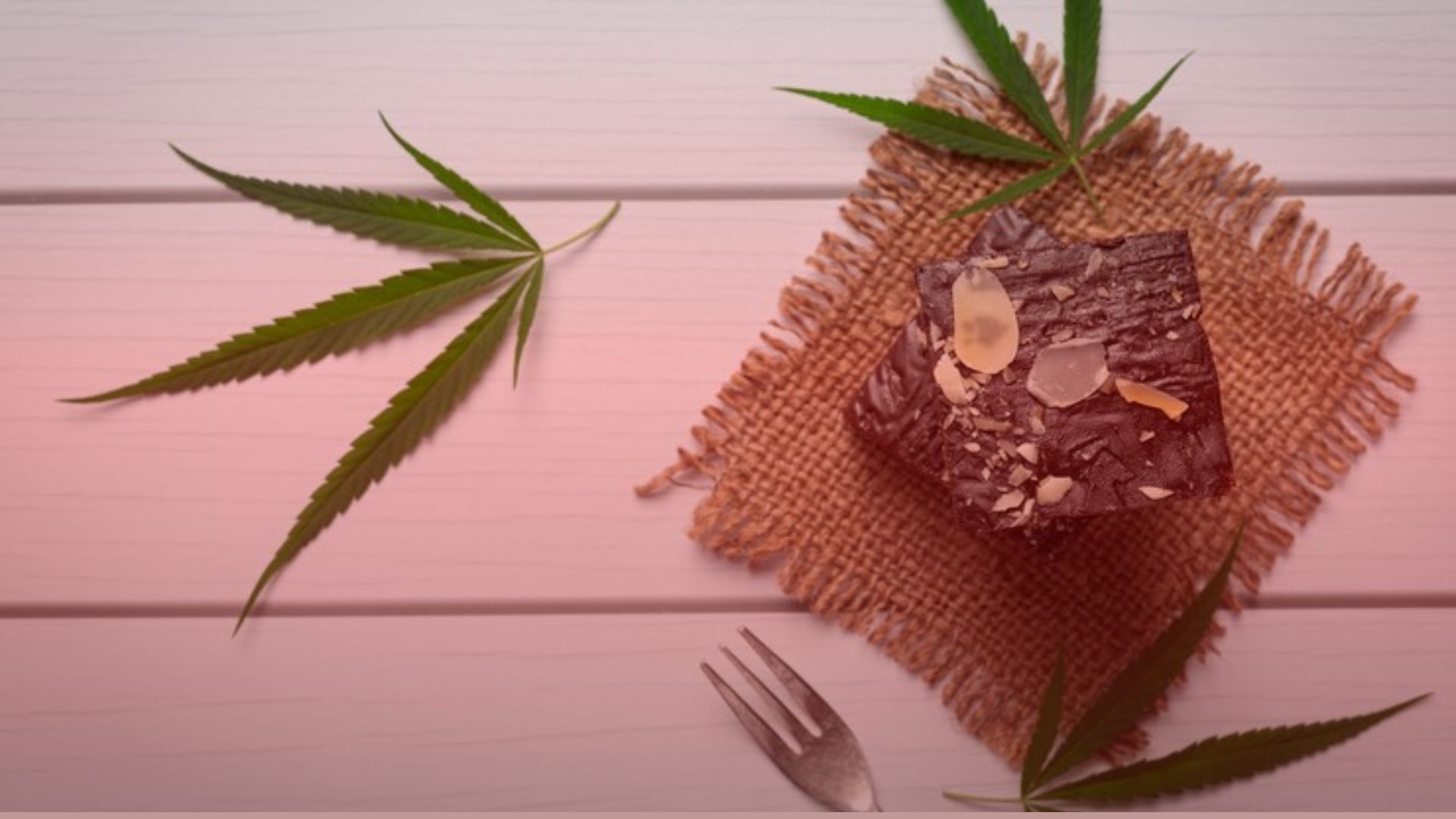 Nature's Canopy House A chocolate brownie with nuts rests on a burlap mat, surrounded by cannabis leaves atop a white wooden surface, with a fork beside it. This delightful treat could be from Natures Canopy House, your go-to dispensary in Mississauga. Dispensary In Mississauga