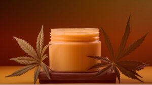 Nature's Canopy House A jar of orange cream sits on a wooden surface, flanked by two large cannabis leaves, evoking the essence of cannabis concentrates against a gradient orange background. Dispensary In Mississauga