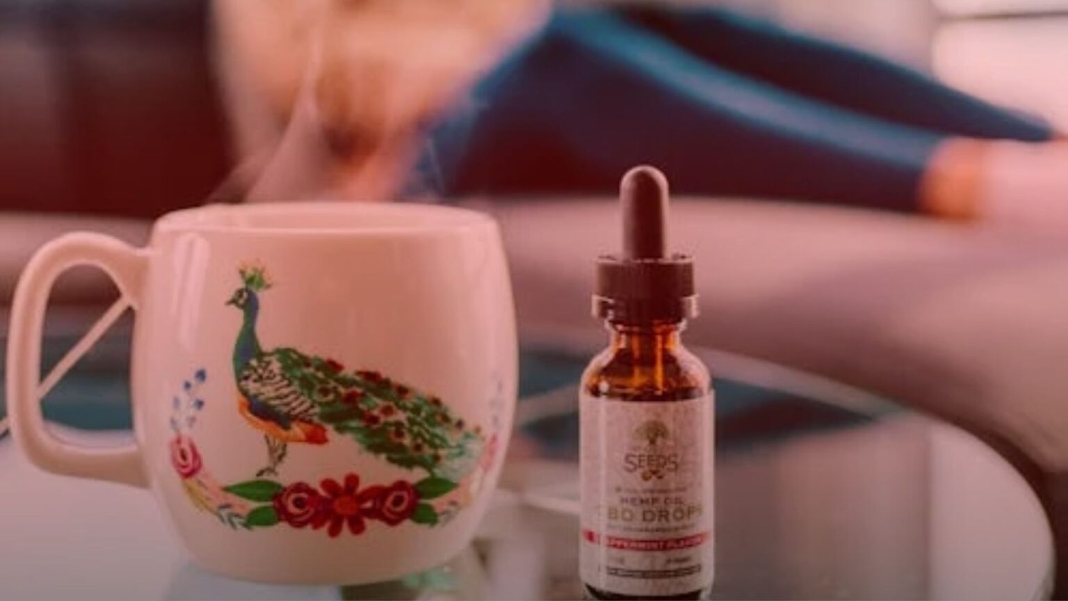 Nature's Canopy House A steaming mug with a peacock design sits next to a bottle labeled "Hemp Oil CBD Drops" on a glass table, perfect for those seeking CBD for stress relief. In the blurred background, a person lounges comfortably, embodying relaxation. Dispensary In Mississauga