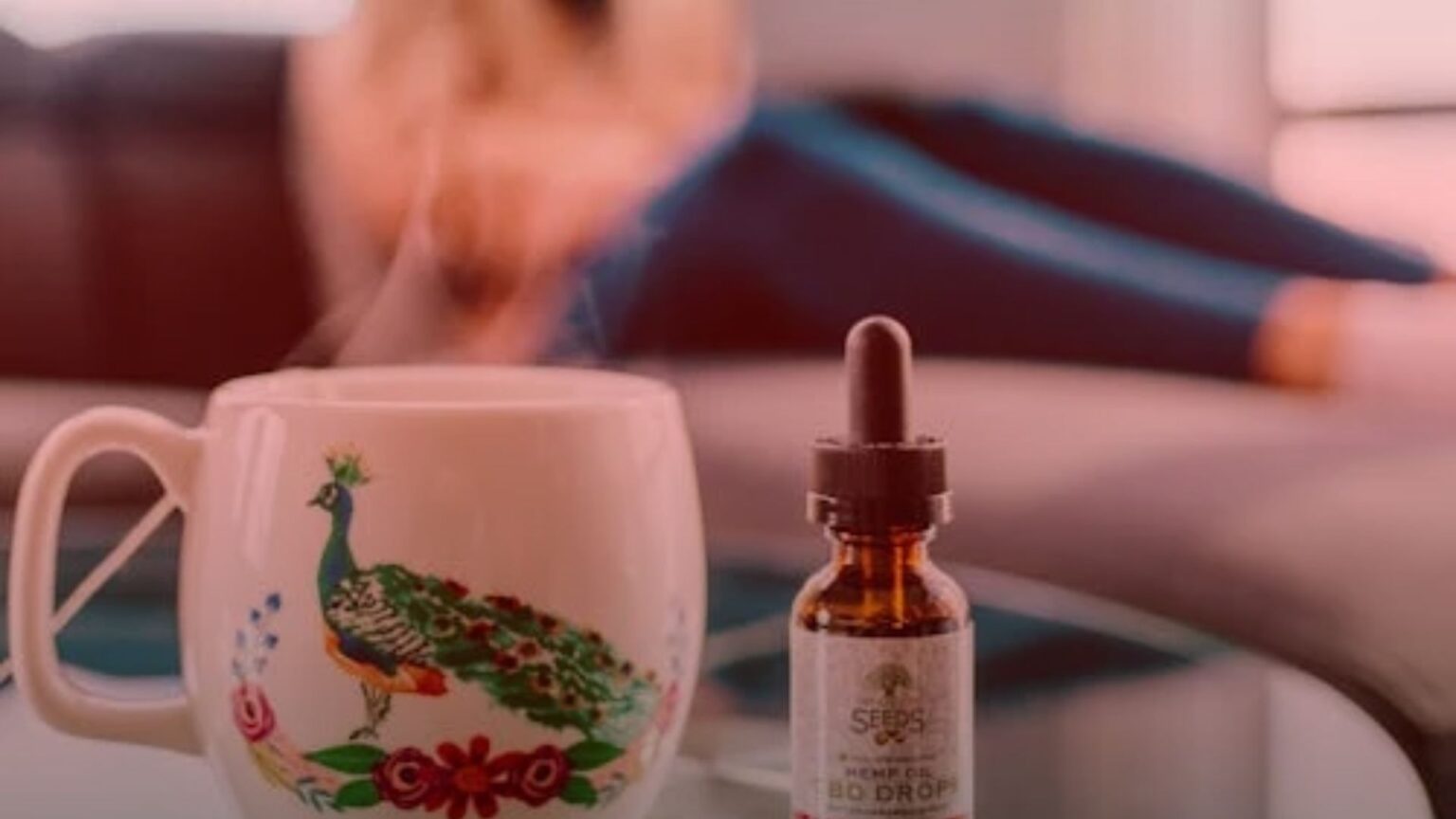 Nature's Canopy House A steaming mug with a peacock design and a bottle of CBD drops, showcasing their healing power, sit on a glass table. A person is blurred in the background. Dispensary In Mississauga